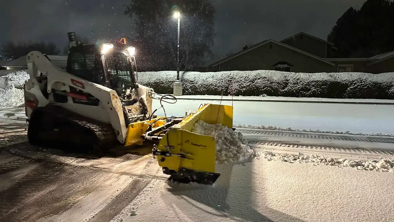 Crystal Clear skid steer with a yellow snow pusher clearing a lot at night
